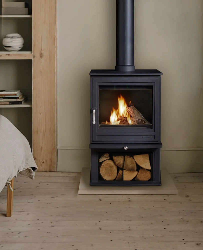 A black wood-burning stove with a glass door, burning firewood inside, set against a beige wall. Below the stove, there is a compartment filled with chopped firewood. To the left, part of a beige armchair and a wooden shelf with books and a ceramic vase are visible.