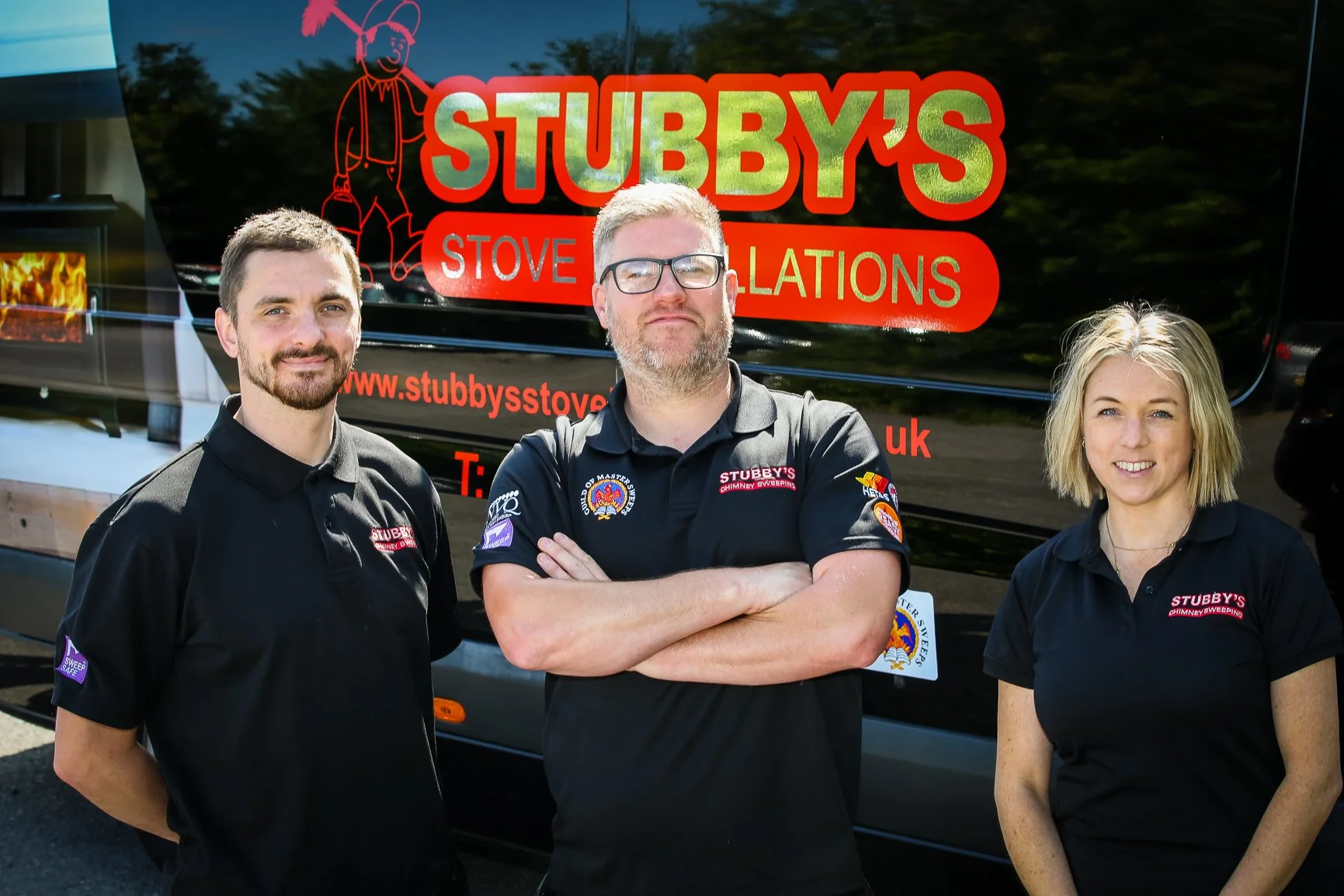 Three people standing in front of a black vehicle with the logo and name of Stubby's Stove Installations. All three are wearing black shirts with company logos. The person in the middle has glasses and is crossing his arms; the person on the left has a beard, and the person on the right has blonde hair. The background shows trees and a clear sky.
