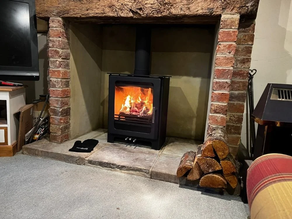 A cozy living room with a fireplace, burning with fire, brick surround, and a wood storage stack to the right. There is a small black object on the stone hearth, a television standing on a white entertainment center to the left, and part of a red and