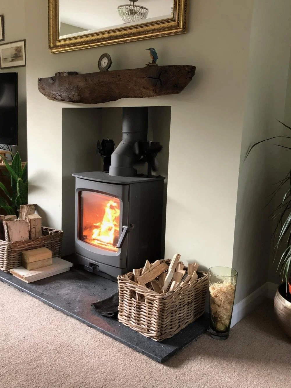 A cozy corner featuring a wood-burning stove with a visible fire, baskets of firewood, a glass vase with wood shavings, a large rustic wooden mantel with decorative items, and a mirror hanging above.