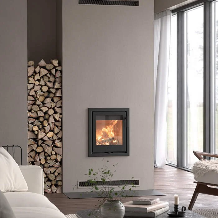 Living room with a modern fireplace, a stack of firewood in a built-in compartment, a white couch, a side table with books and a plant, and large sliding glass doors with white curtains.