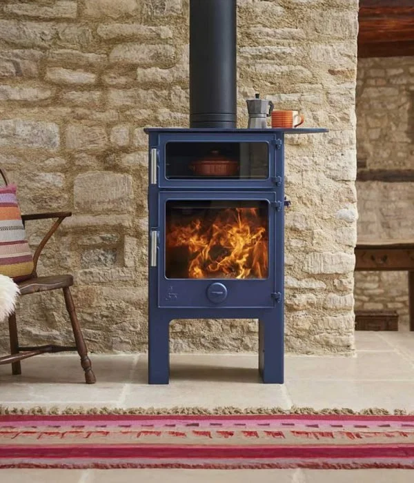 Blue wood-burning stove with visible flames inside, placed against a brick wall with a shelf on top holding a pitcher and cups.