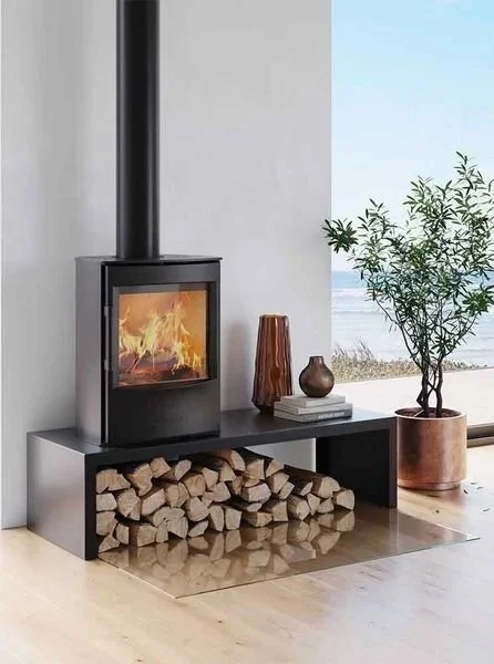 Modern indoor fireplace with a wood stack underneath, decorated with vases and a potted plant, near a window overlooking a body of water.