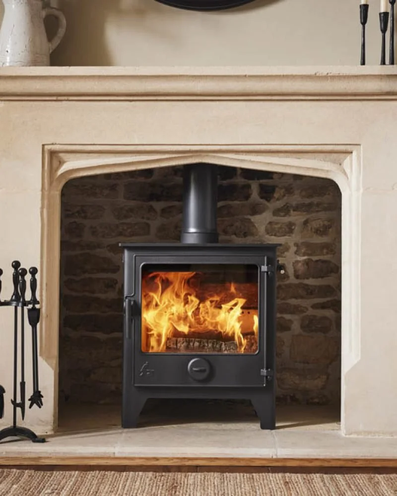 A wood-burning stove with visible flames inside, set in a stone fireplace with a carvedstone surround, located in a cozy living room.