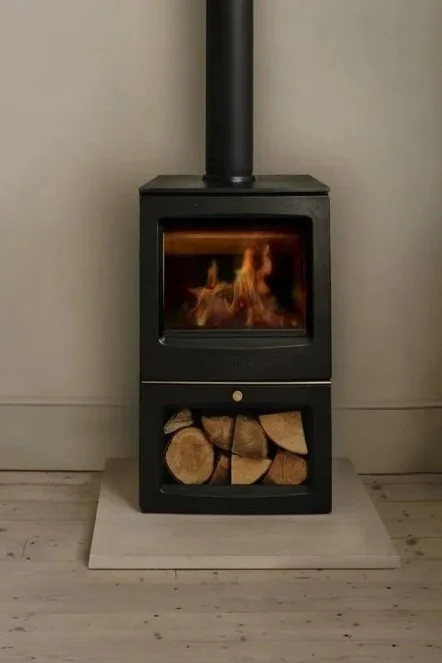 A black wood stove with a glass door showing a fire inside, placed on a white hearth with firewood stored below, against a plain wall.