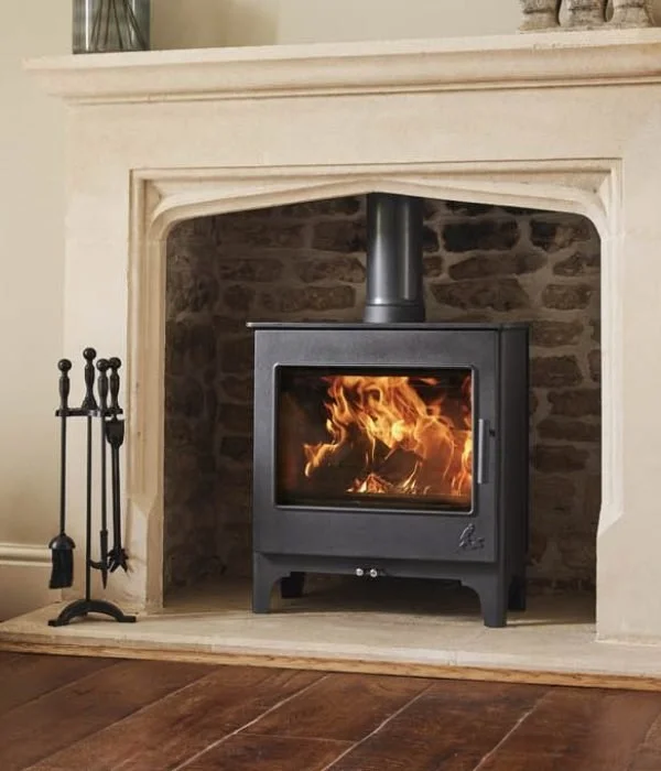 A black wood stove burning with flames inside a fireplace with stone interior, surrounded by a cream-colored mantel and hearth in a living room.