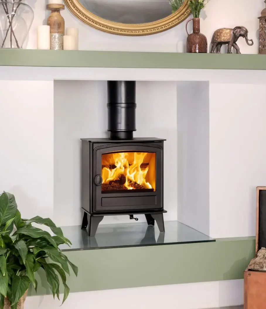 A black wood stove with a visible fire inside, sitting on a hearth within a white alcove in a living room. The alcove has a green shelf above it, decorated with candles, vases, and a decorative elephant sculpture. A large mirror hangs above the shelf, and a green leafy plant is partially visible on the left side.