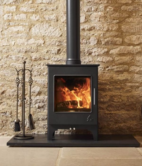A modern wood-burning stove with a fire inside, next to a metal fireplace toolset, against a textured stone wall.