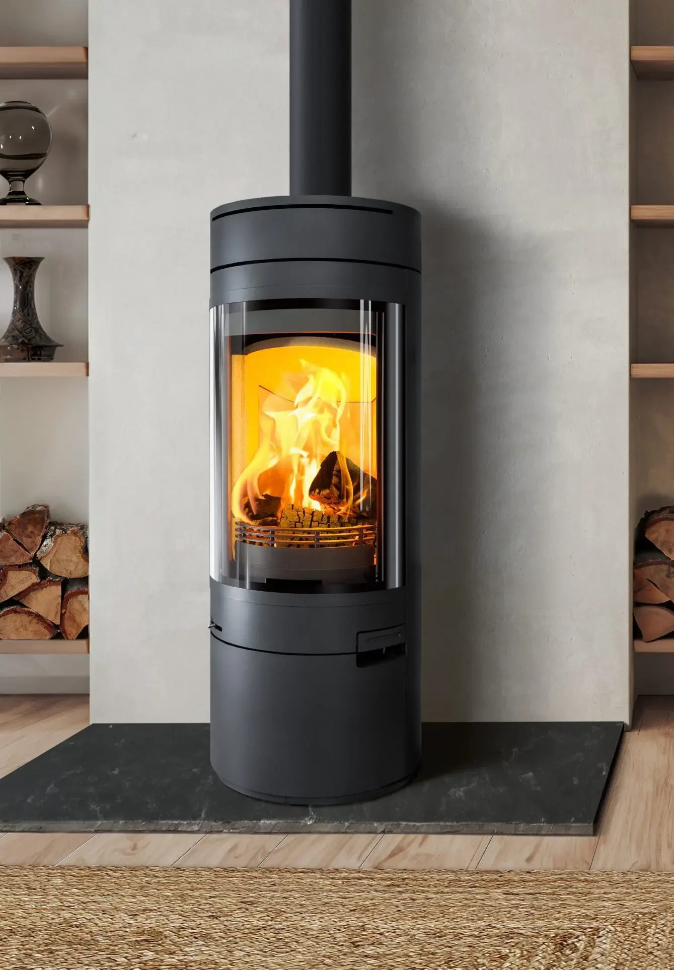 A modern cylindrical black pellet stove with a glass front showing flames inside, positioned on a black platform in a living room with wood flooring and built-in shelves on each side.