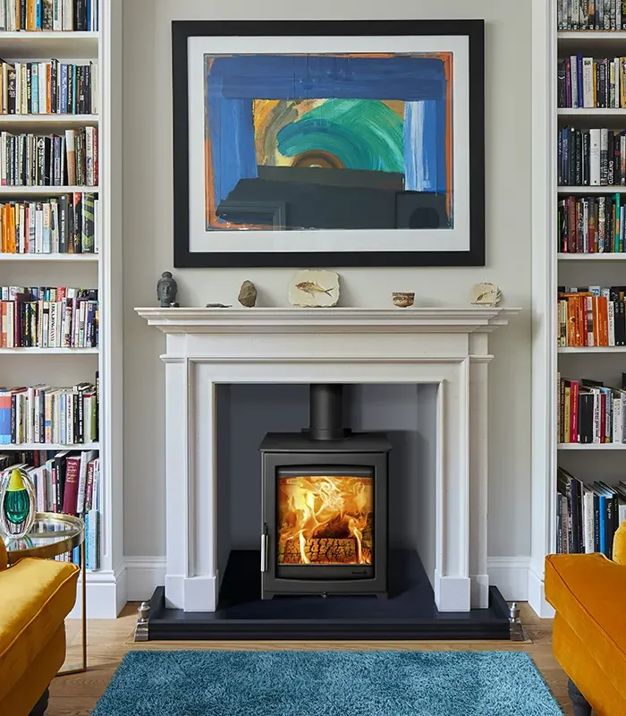 A cozy living room with a white fireplace, a wood-burning stove with fire inside, surrounded by bookshelves filled with books, a colorful abstract painting above the fireplace, and two mustard-colored armchairs on either side.