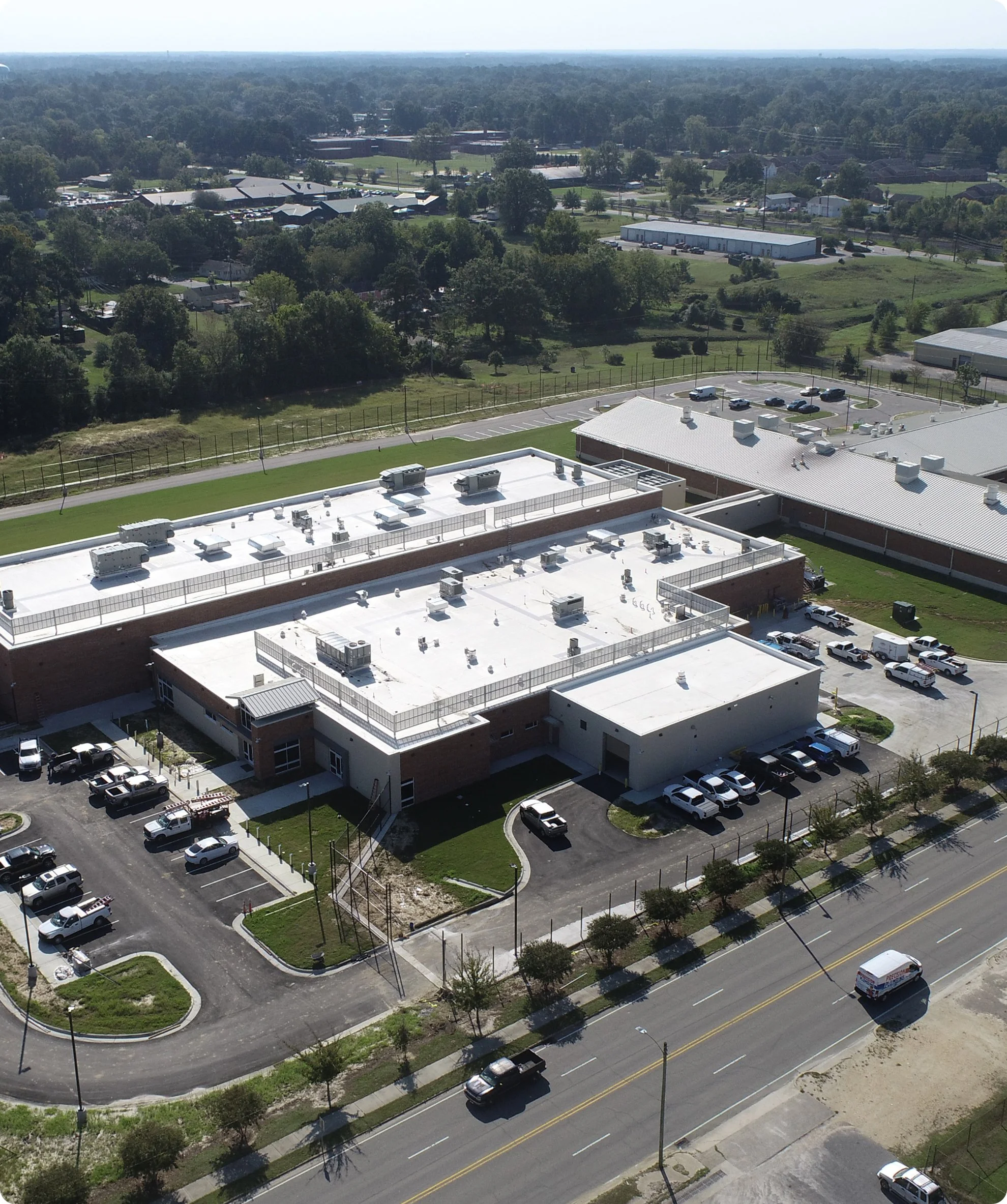 An aerial view of a commercial and residential area with parking lots, buildings, trees, and a two-lane road with vehicles.