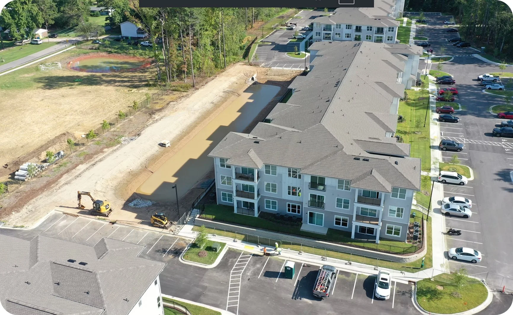 An aerial view of an apartment complex under construction with ongoing groundwork and landscaping. There are construction vehicles and equipment on the site, and an adjacent parking lot with parked cars.