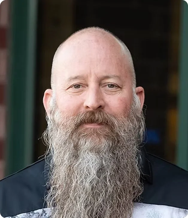 A middle-aged man with a long gray beard, bald head, and light skin, wearing a dark jacket and standing outdoors.