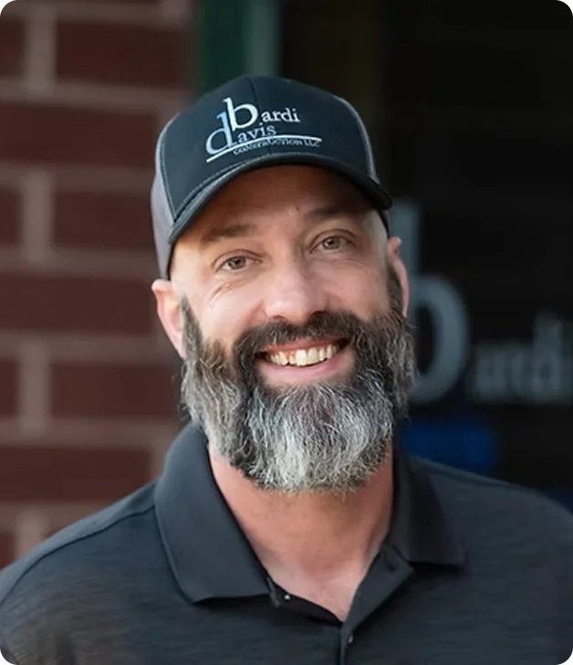 A smiling man with a beard and mustache wearing a black cap and black polo shirt, standing outdoors in front of a brick wall.