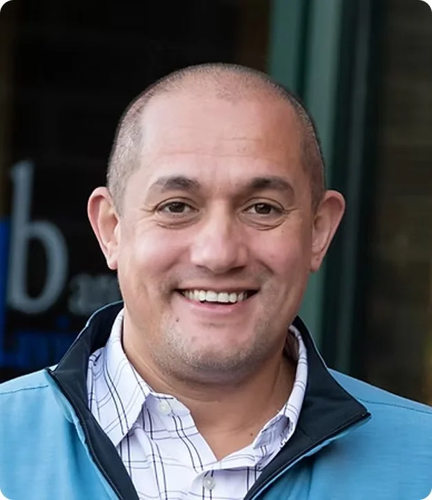 Smiling man with short shaved hair wearing a collared shirt and blue jacket, standing outdoors.