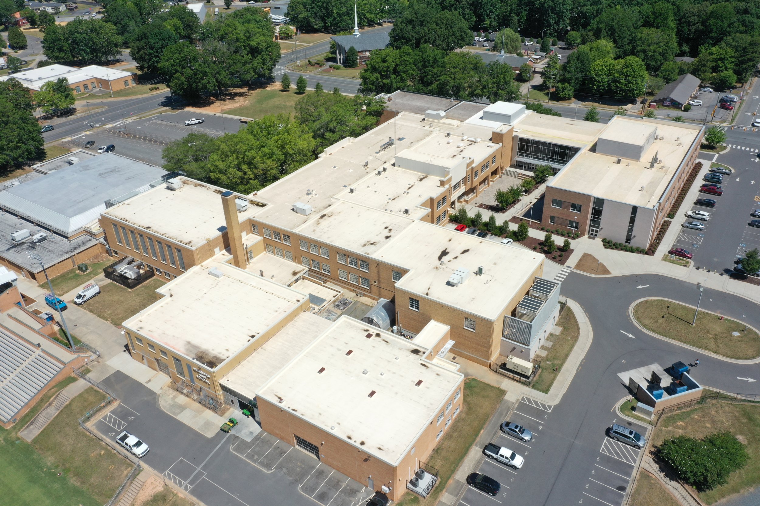 Aerial view of a large school building with a parking lot, surrounded by trees, roads, and smaller buildings.
