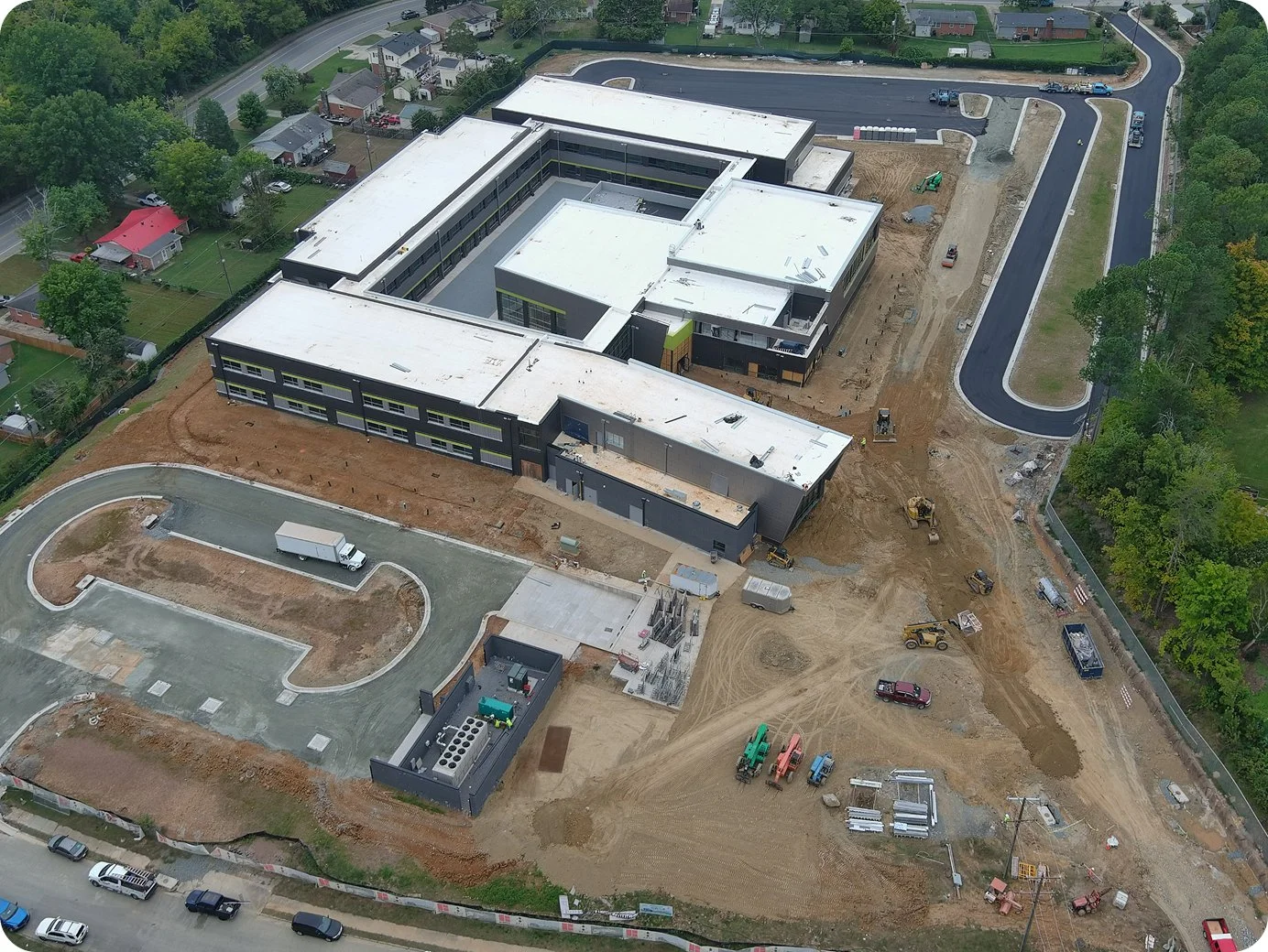 An aerial view of a large building under construction, with a curved road in the front and a rectangular parking lot in the back. Construction vehicles and dirt areas are visible around the site.