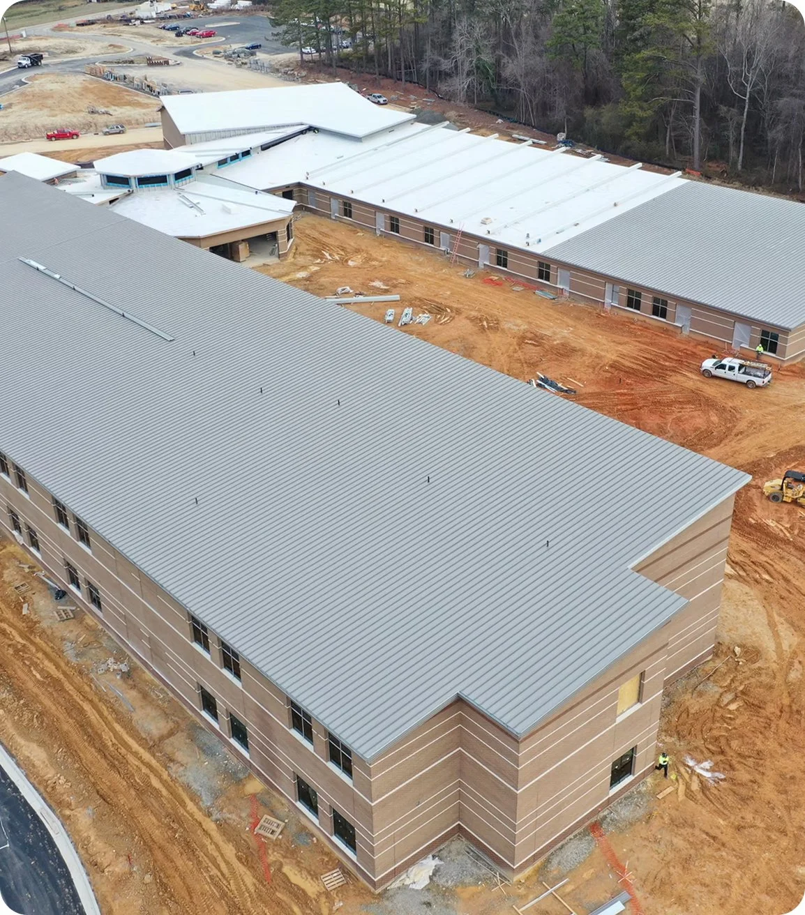 An aerial view of a construction site showing multiple buildings in progress with metal roofs, dirt ground, construction vehicles, and workers.
