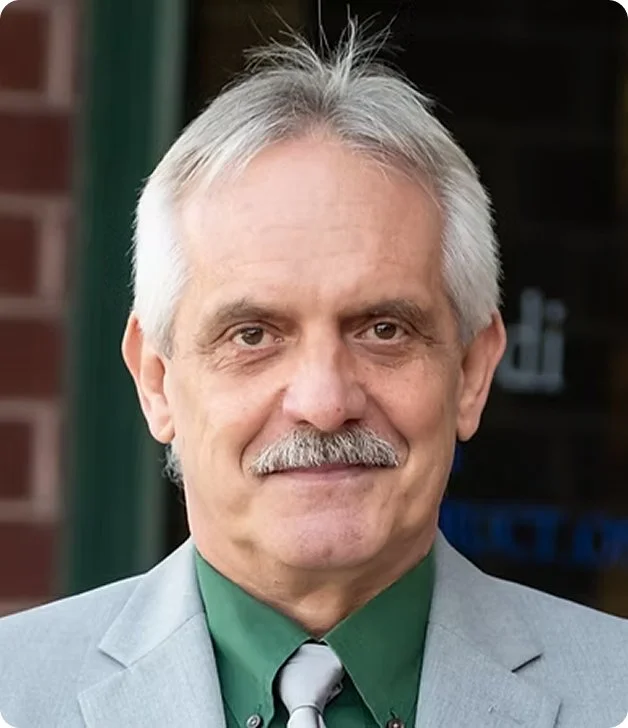 A middle-aged man with gray hair, a mustache, and a friendly expression, wearing a light gray suit, green shirt, and a gray tie, standing outside near a building.