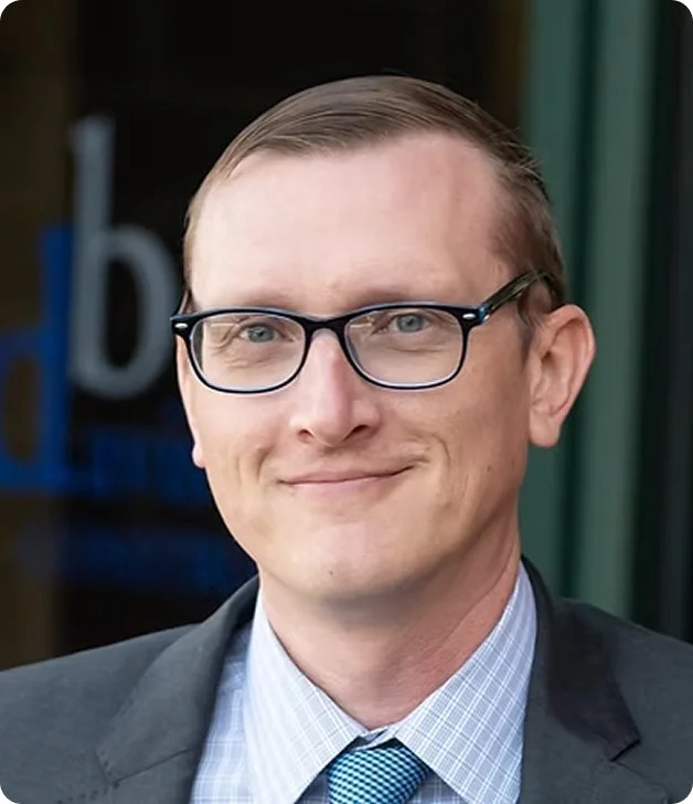 A man with short brown hair, wearing glasses, a dark suit jacket, a light blue checkered shirt, and a blue tie, smiling outdoors.