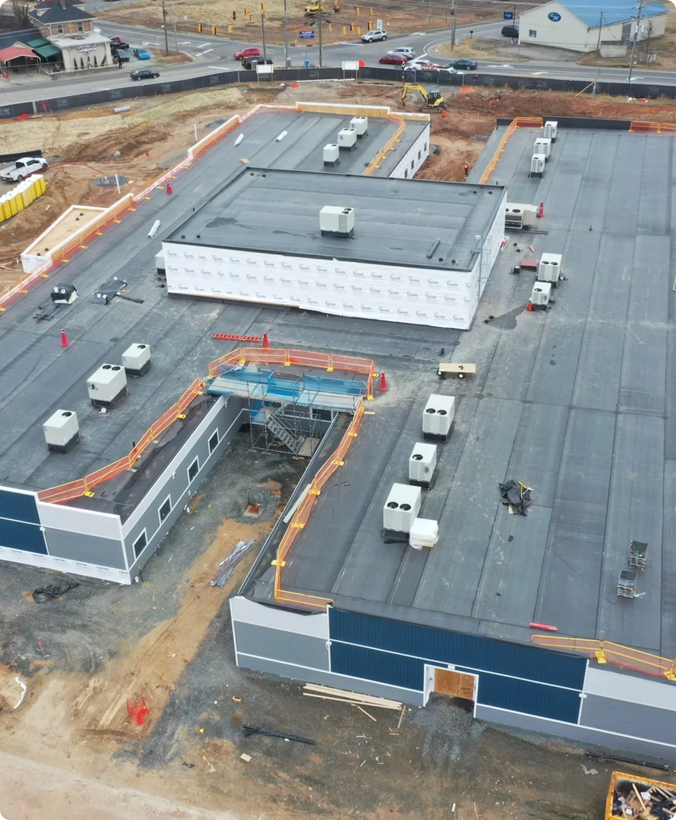 Aerial view of a building under construction with a flat roof, HVAC units, construction equipment, orange safety fencing, and surrounding dirt and pavement, near a busy road with vehicles.