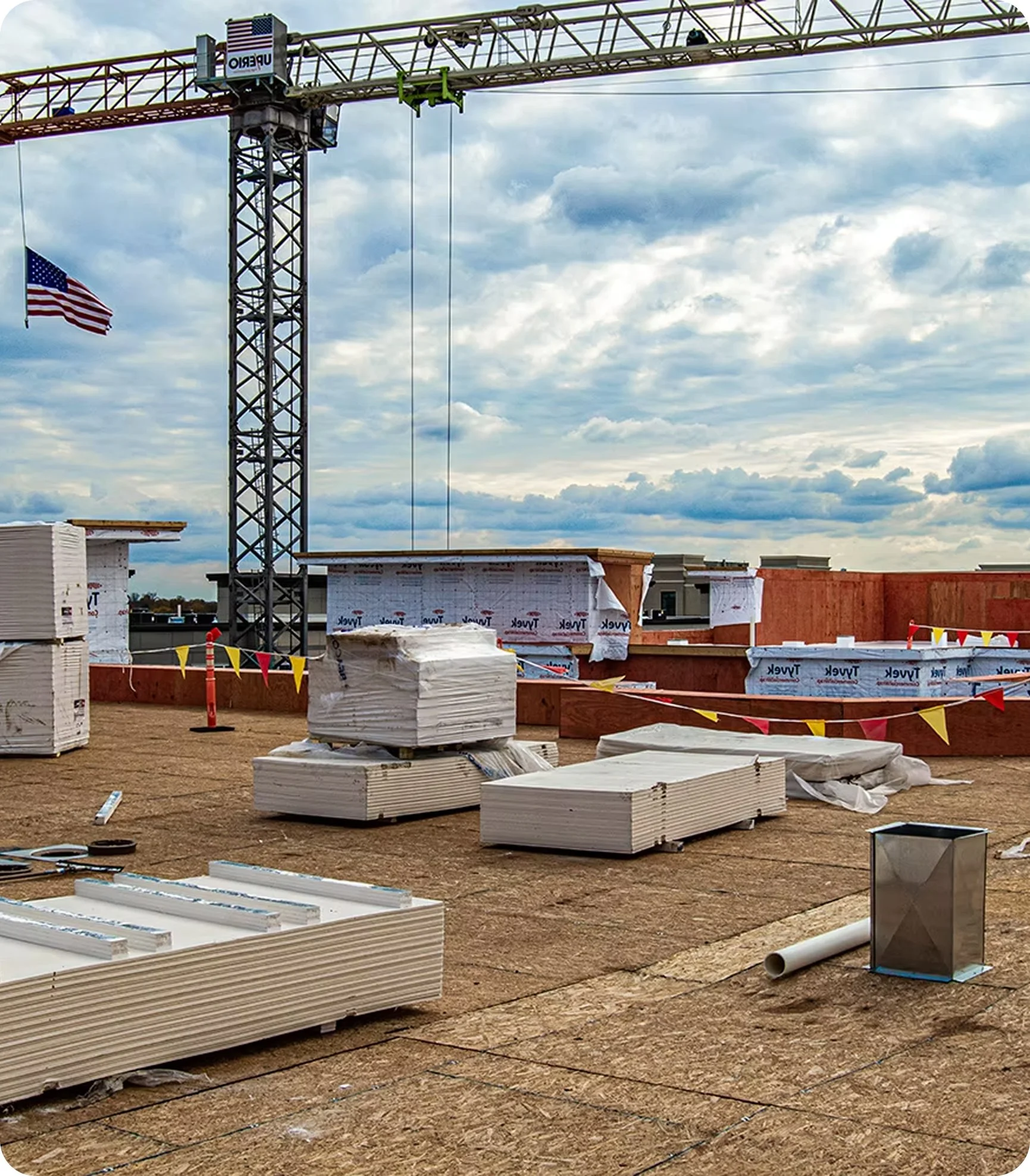 Construction site with building materials, a crane, and an American flag under a cloudy sky.