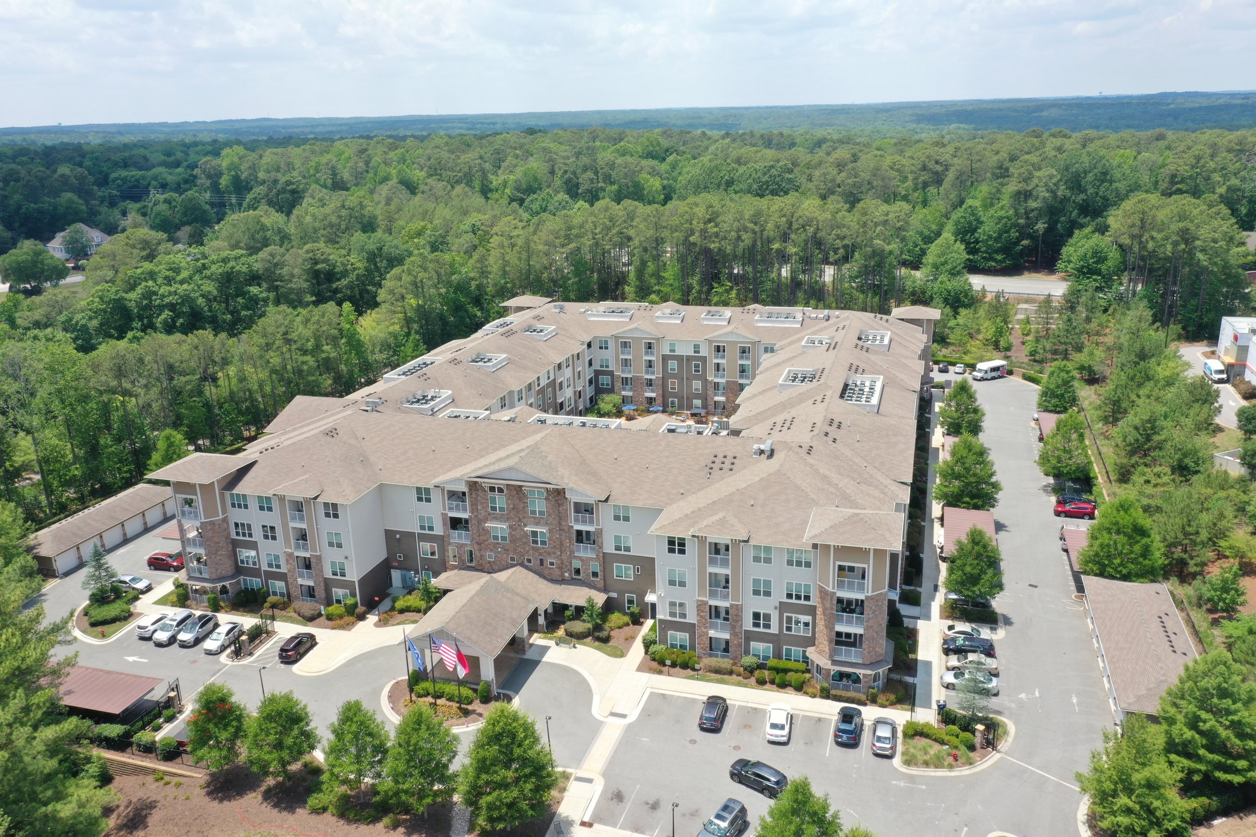 An aerial view of a multi-story residential building surrounded by green trees, with parking lots and cars, set in a lush wooded area extending to the horizon under a partly cloudy sky.