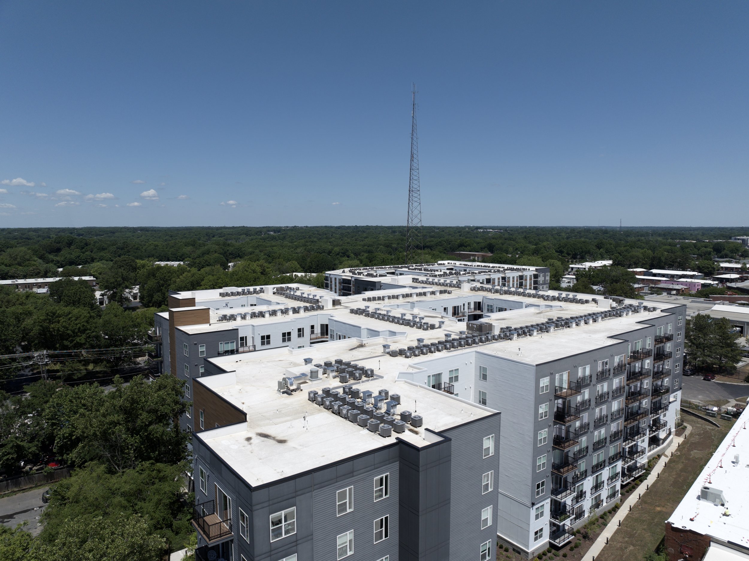 Aerial view of a cityscape with modern white apartment buildings with rooftop HVAC units, surrounded by lush green trees and a radio tower in the distance under a clear blue sky.