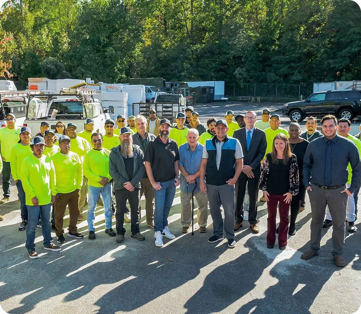 A large group of people, some wearing neon yellow shirts and caps, gathered outdoors on a paved area, with some men in casual and business attire. The background shows parked trucks, trailers, and a chain-link fence with trees behind.