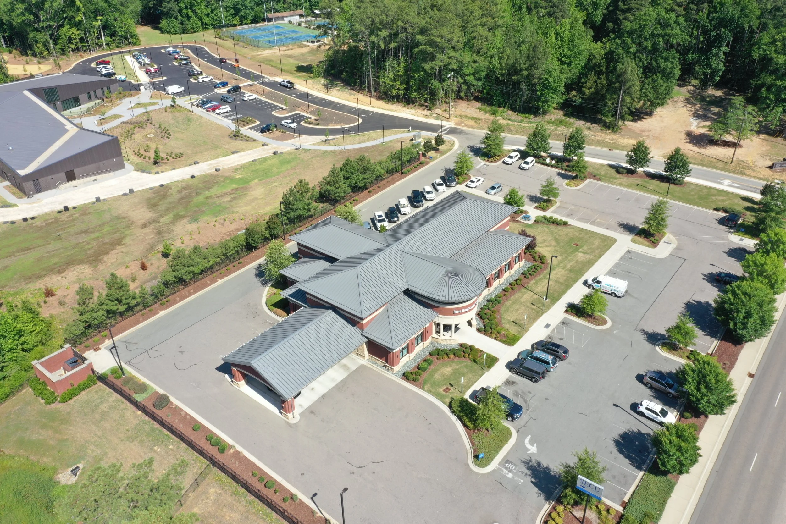 Aerial view of a parking lot next to a building with a gray roof, surrounded by trees and greenery, with some parked cars, a tennis court, and a community area.