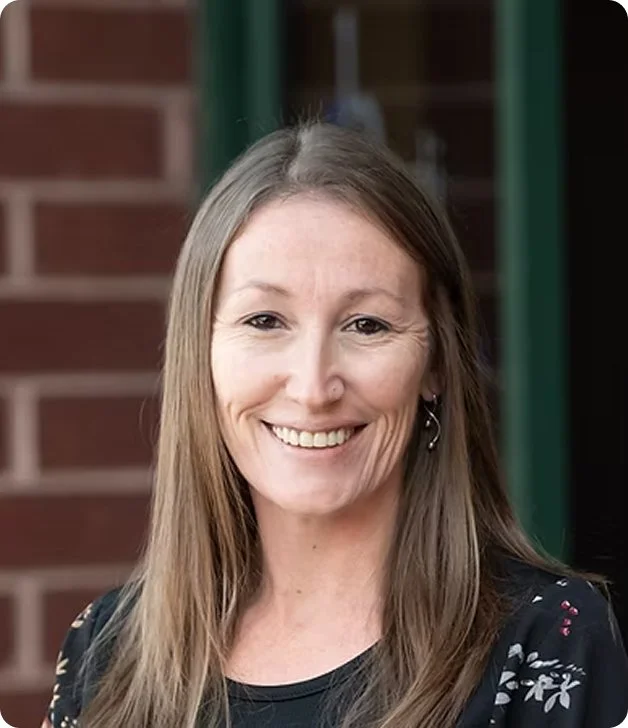 A woman with light brown hair smiling outdoors, with a brick wall and green pole in the background.