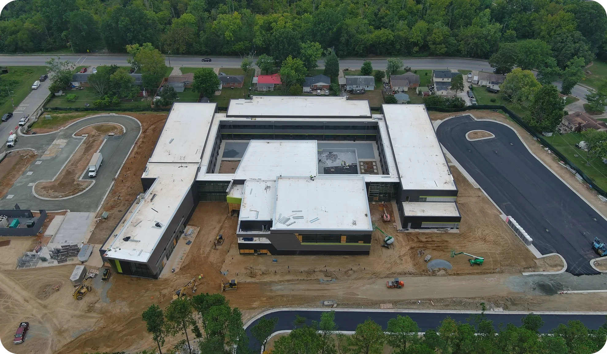 An aerial view of a large building under construction with a parking lot and surrounding streets, with trees and residential houses in the background.