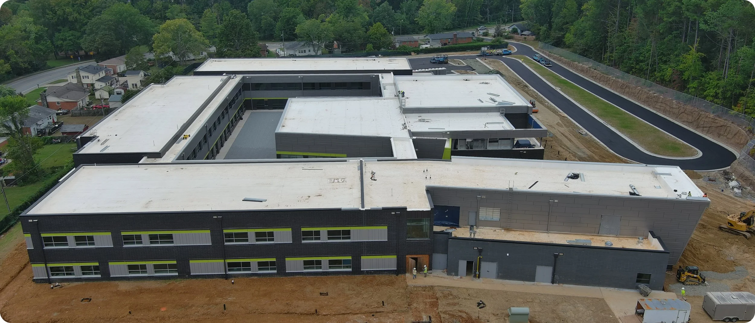 Aerial view of a modern school building under construction with a curved asphalt driveway and surrounding greenery.