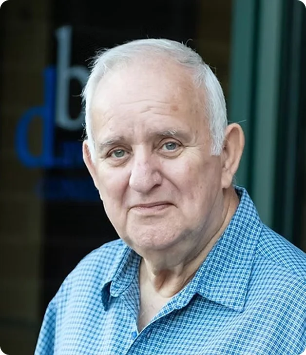 An elderly man with white hair and blue eyes wearing a blue checkered shirt, standing outdoors.