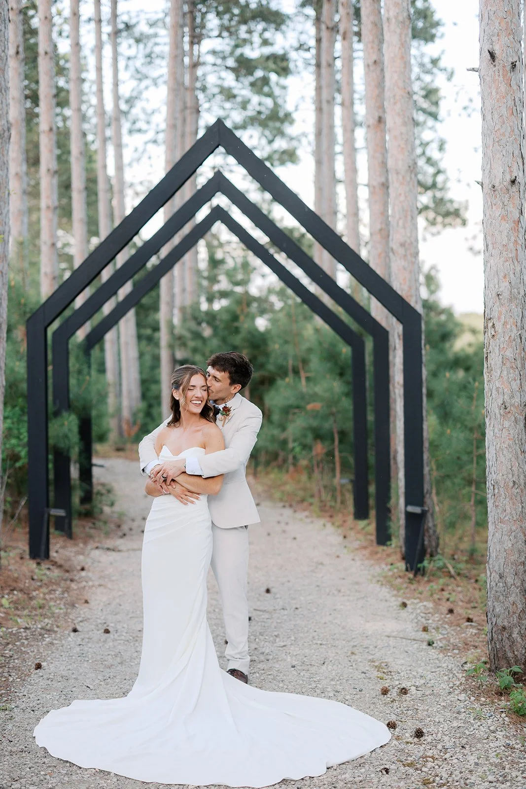 A newlywed couple in white wedding attire standing on a forest trail beneath a black geometric arch, surrounded by tall trees and greenery.