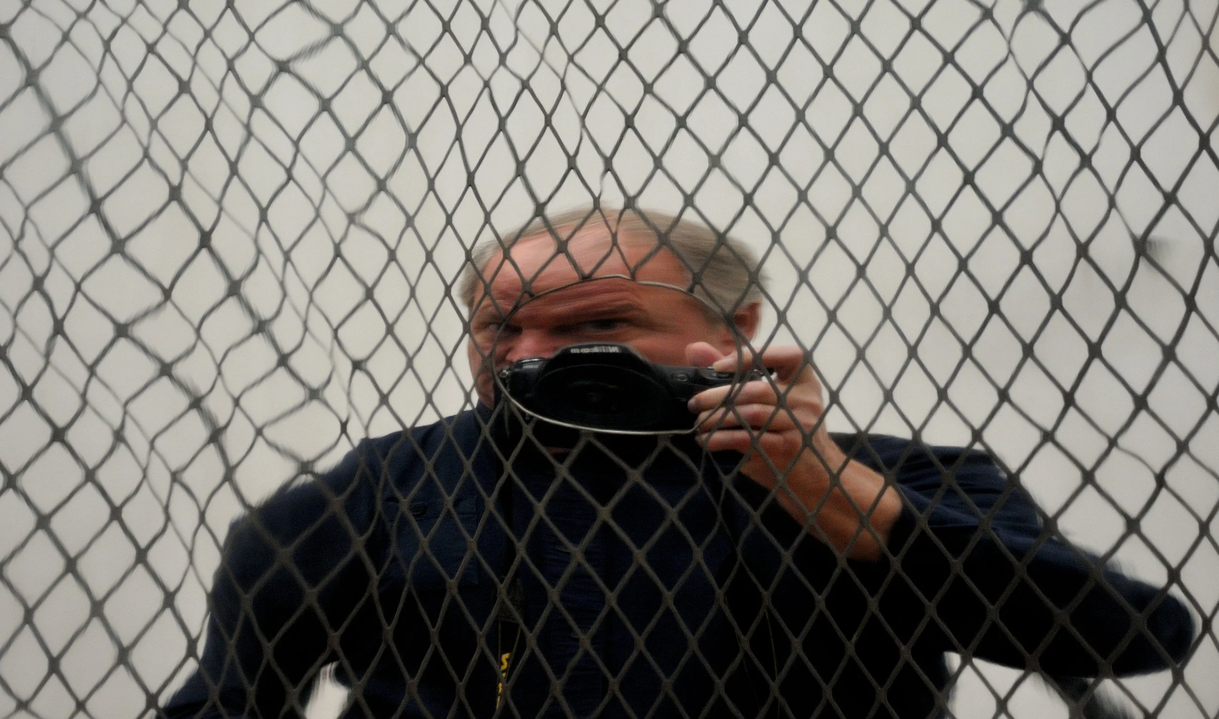 A person taking a selfie in front of a mirror, with a metal chain-link fence in front of the mirror, in a room with gray walls.