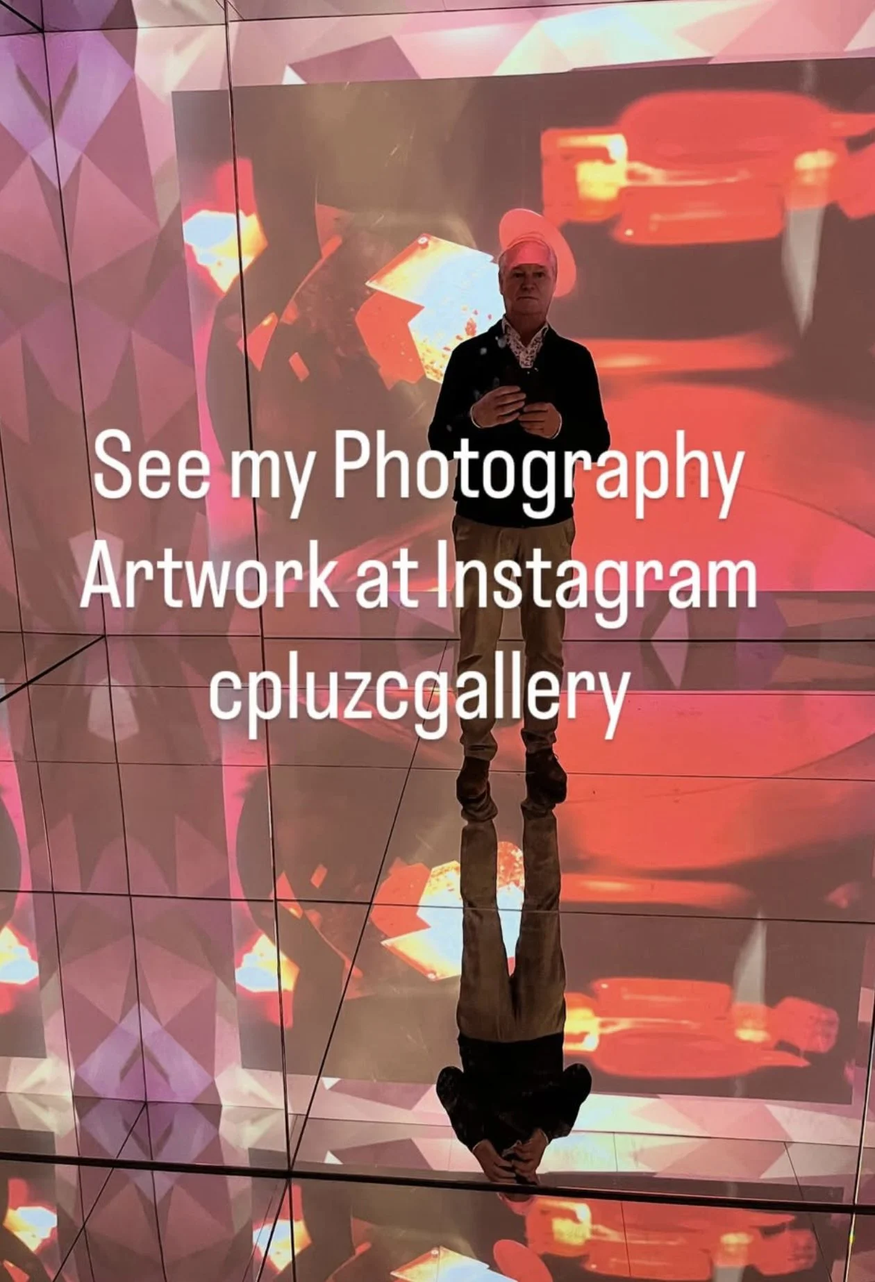 A person wearing a white headscarf, dark jacket, and light-colored pants is standing inside a modern indoor space with large, colorful digital screens or projections on the walls. The individual is holding a phone and is reflected on the shiny, mirror-like floor. Overlaid text reads 'See my Photography Artwork at Instagram cpluzggallery.'