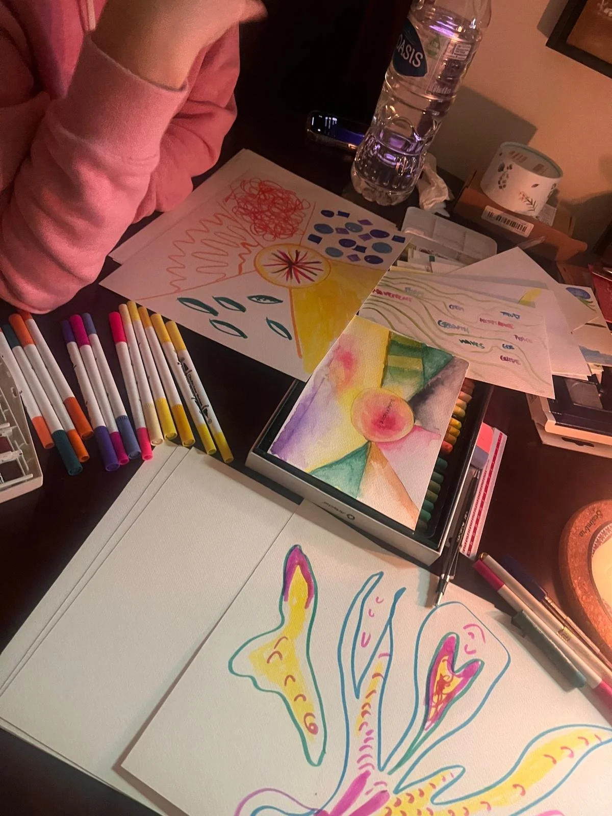 Table with colorful drawings and markers, water bottle, tape, and art supplies. A person wearing a pink hoodie is working on the art projects.