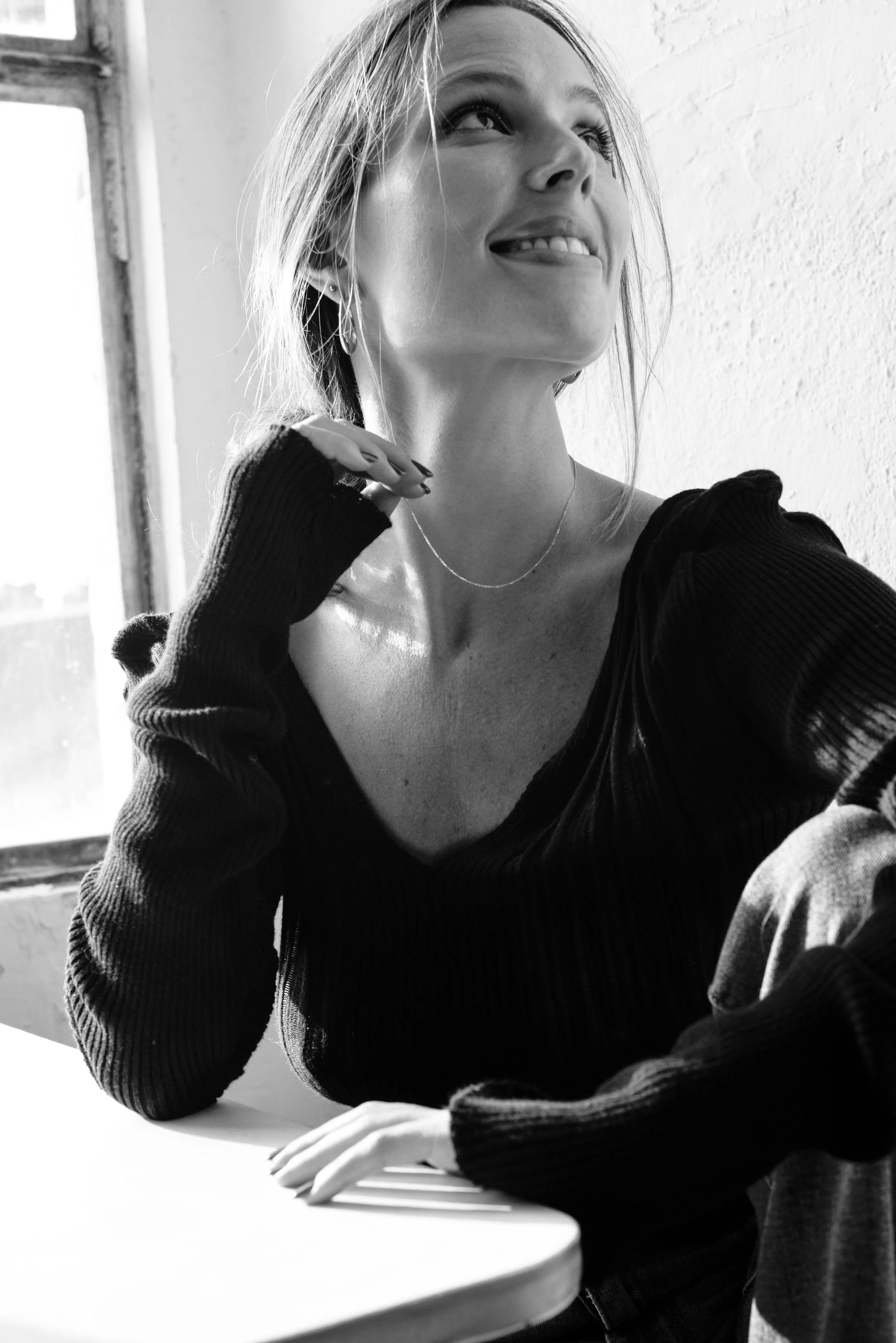 A young woman with light-colored hair, wearing a black top with a zipper, sitting at a table with a relaxed expression. She is gently biting her tongue and looking upward, with her right hand resting on the table and her left hand near her neck, wearing hoop earrings and a delicate chain necklace. The background shows a window with sunlight streaming in.