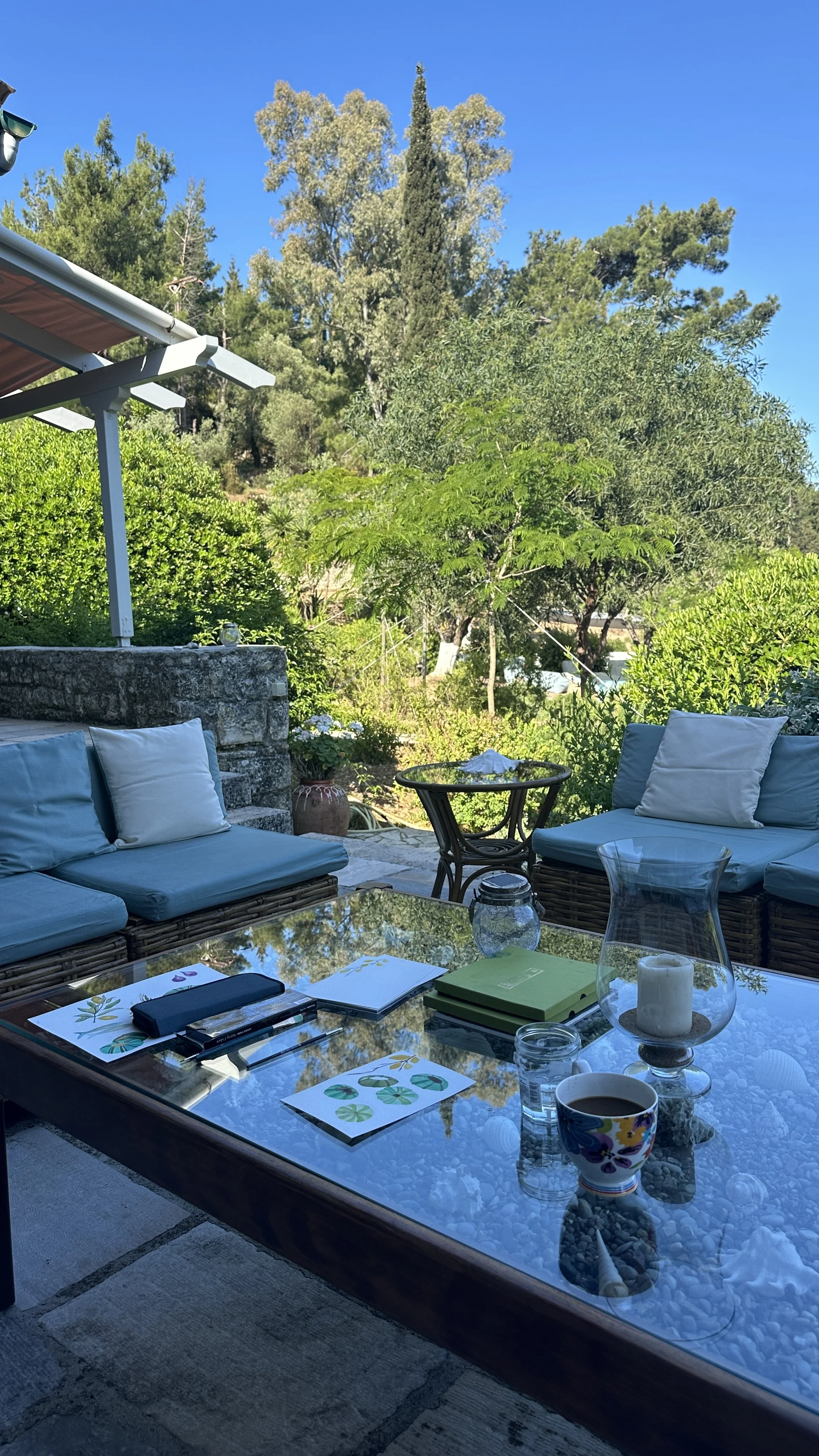 Outdoor patio table with cups, notebooks, and decorative items; surrounded by cushioned seating and lush green trees against a clear blue sky.
