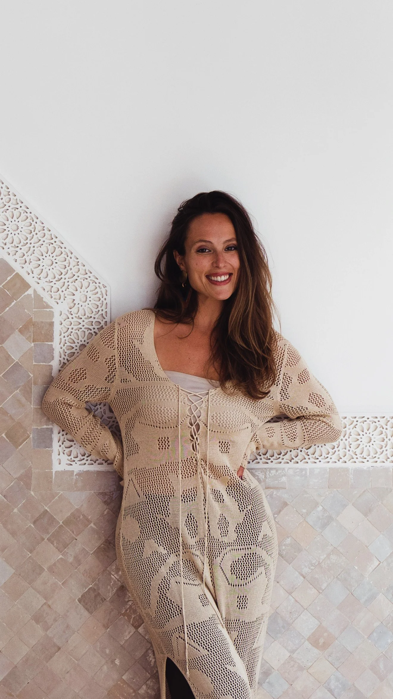 A woman standing indoors against a decorative wall, smiling with her hands on her hips, wearing a light beige crocheted dress.