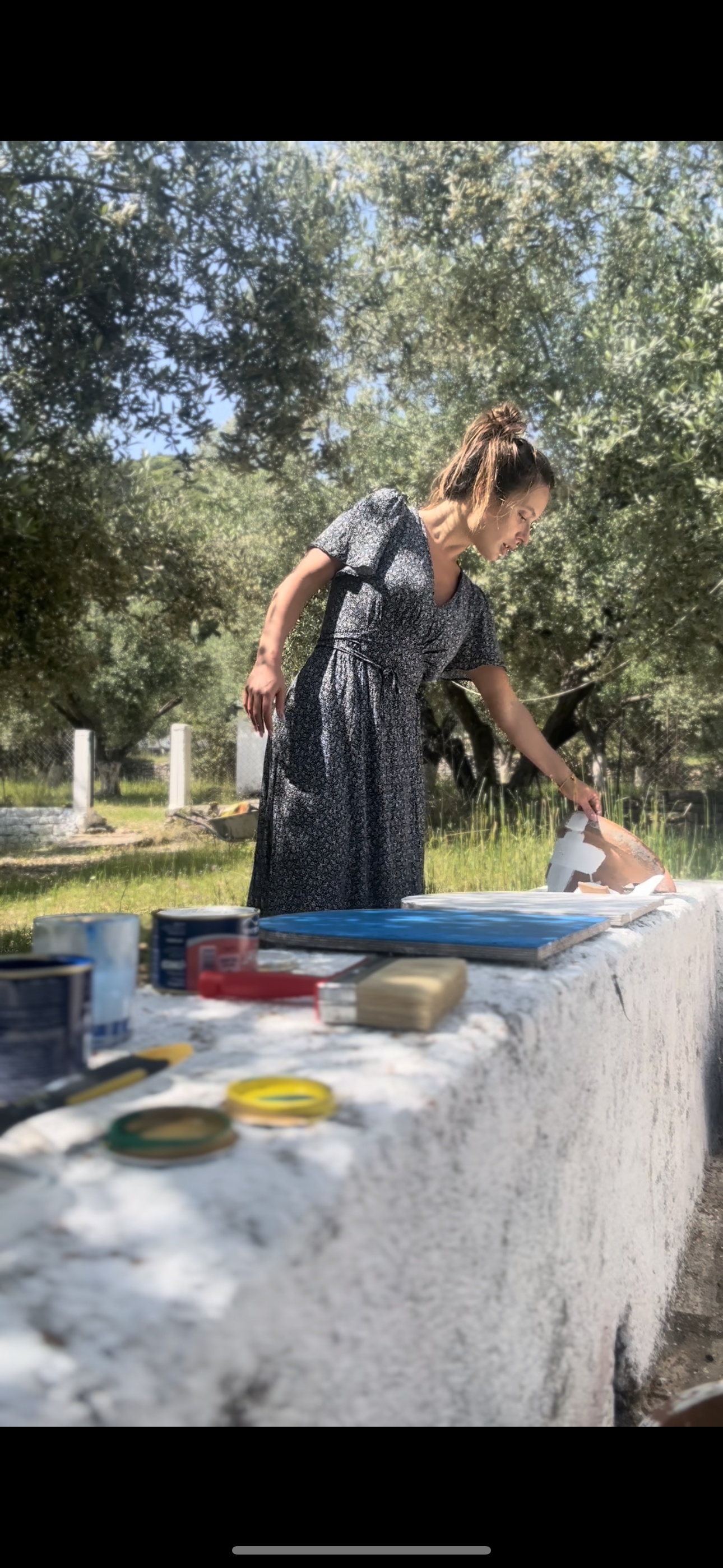 A woman in a black floral dress with a tied waist is standing outdoors under a tree, reaching to interact with a ceramic object on a white stone table. Gardening tools, cans, and brushes are on the table, with a lush garden in the background.