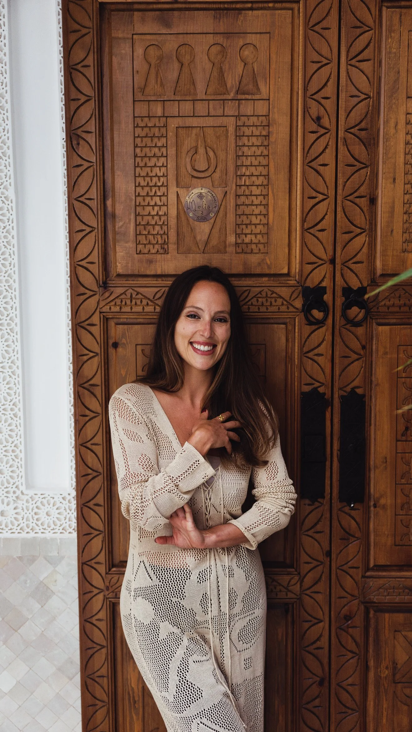 A woman with long brown hair, smiling, wearing a beige, lacy, patterned dress, standing in front of a large wooden carved door.