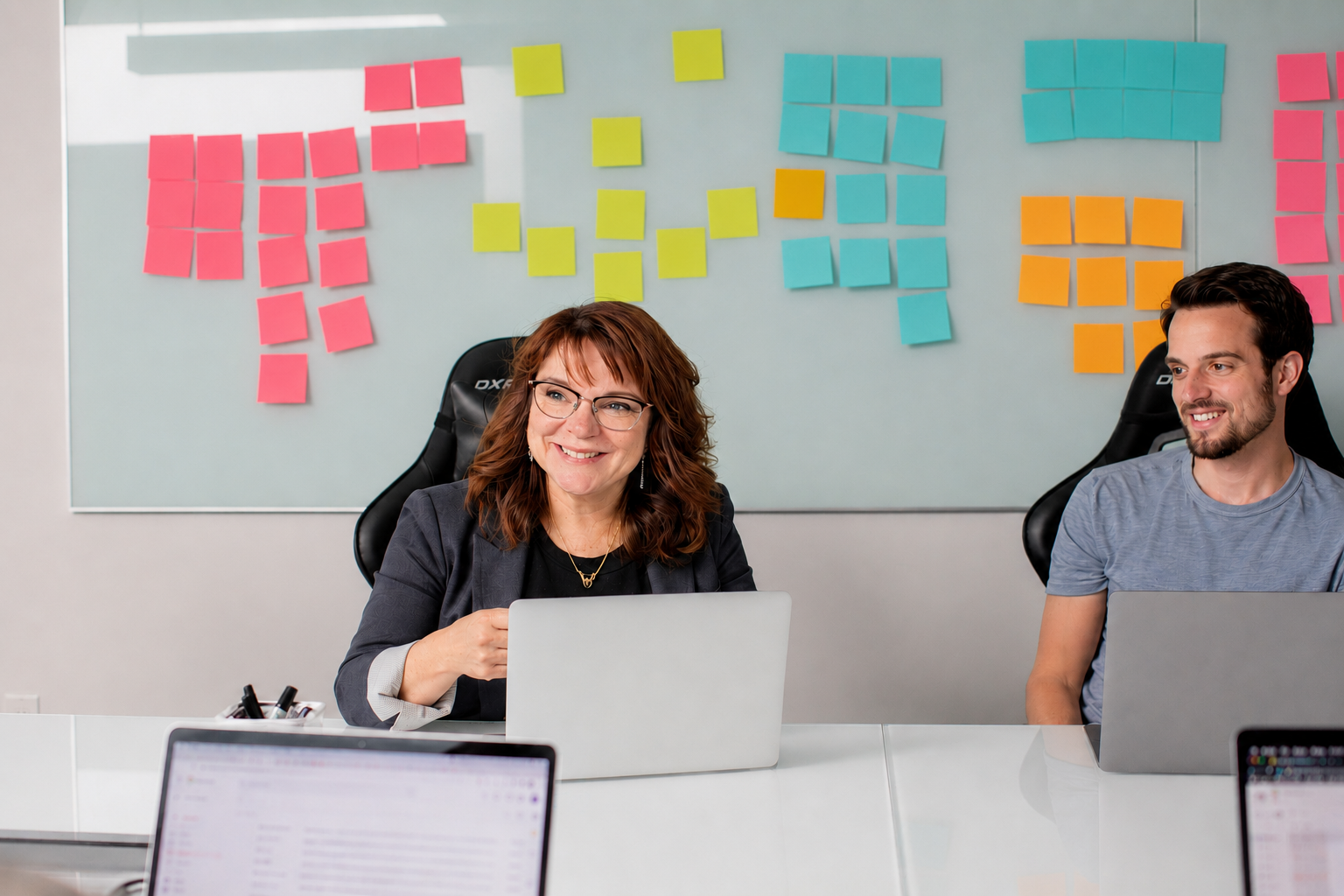 Two people sitting at a conference table in an office, smiling and working on laptops, with colorful sticky notes on a whiteboard behind them.