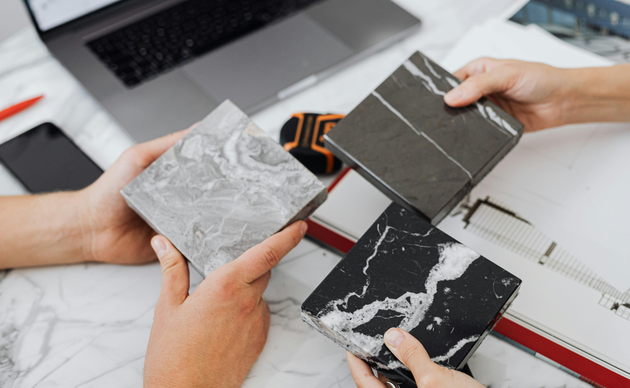 Two people holding sample black and gray marble tiles with white veining, in a workspace with a laptop and tools.