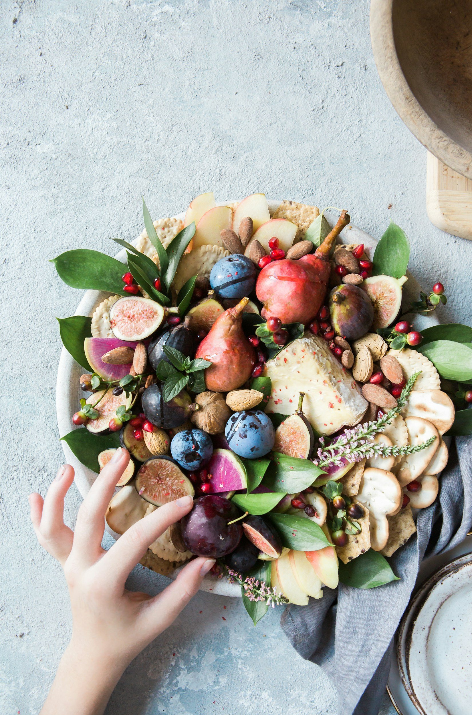 Una gran bandeja redonda con variedad de frutas, nueces y hojas verdes, decorada con flores pequeñas y un peinado de mano en la esquina inferior izquierda.