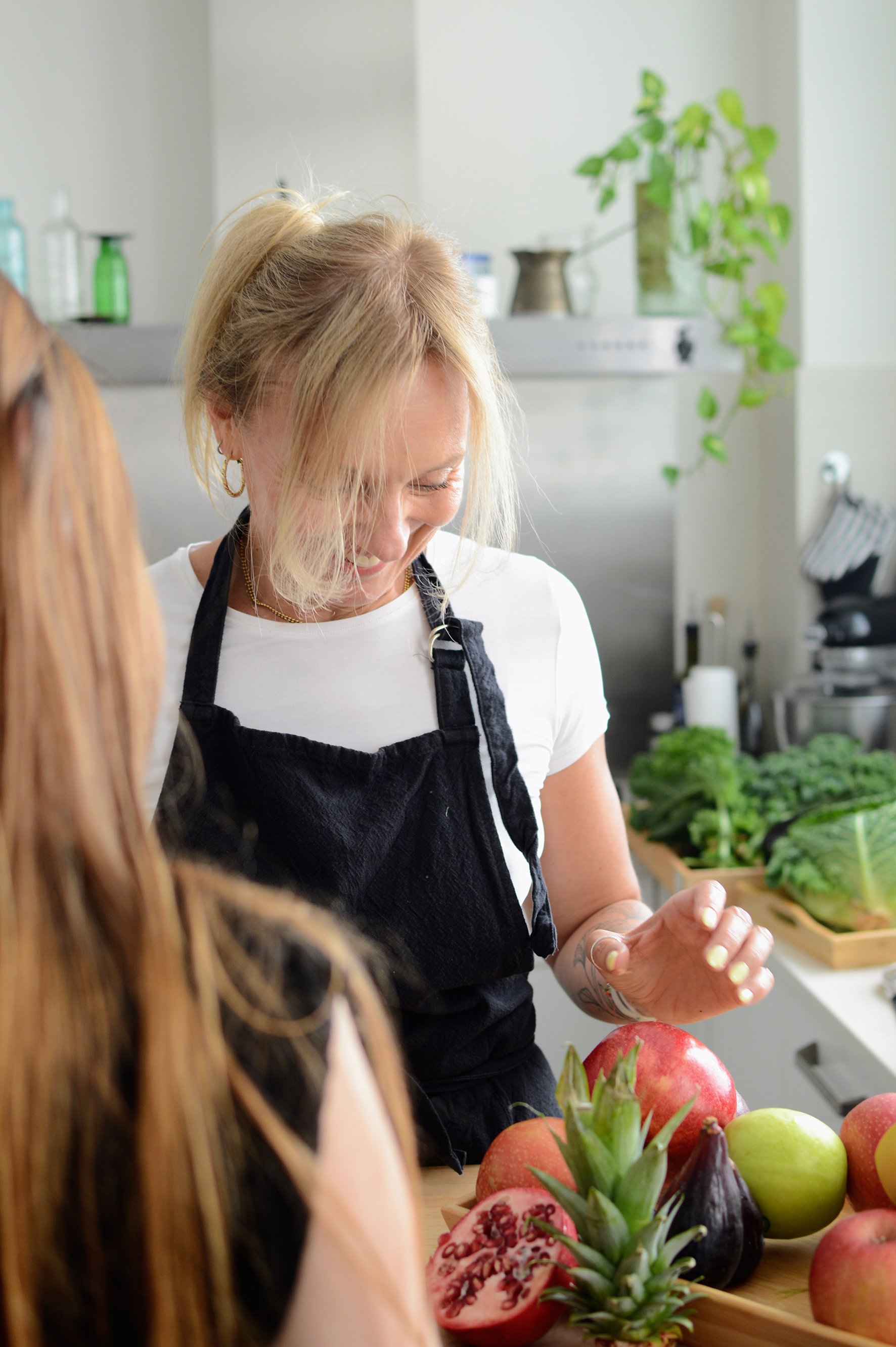 Pepa Poduje, mientras conversa con otra persona cerca de una mesa con frutas, incluyendo granadas, piñas, manzanas y uvas, en una cocina moderna con plantas en el fondo.