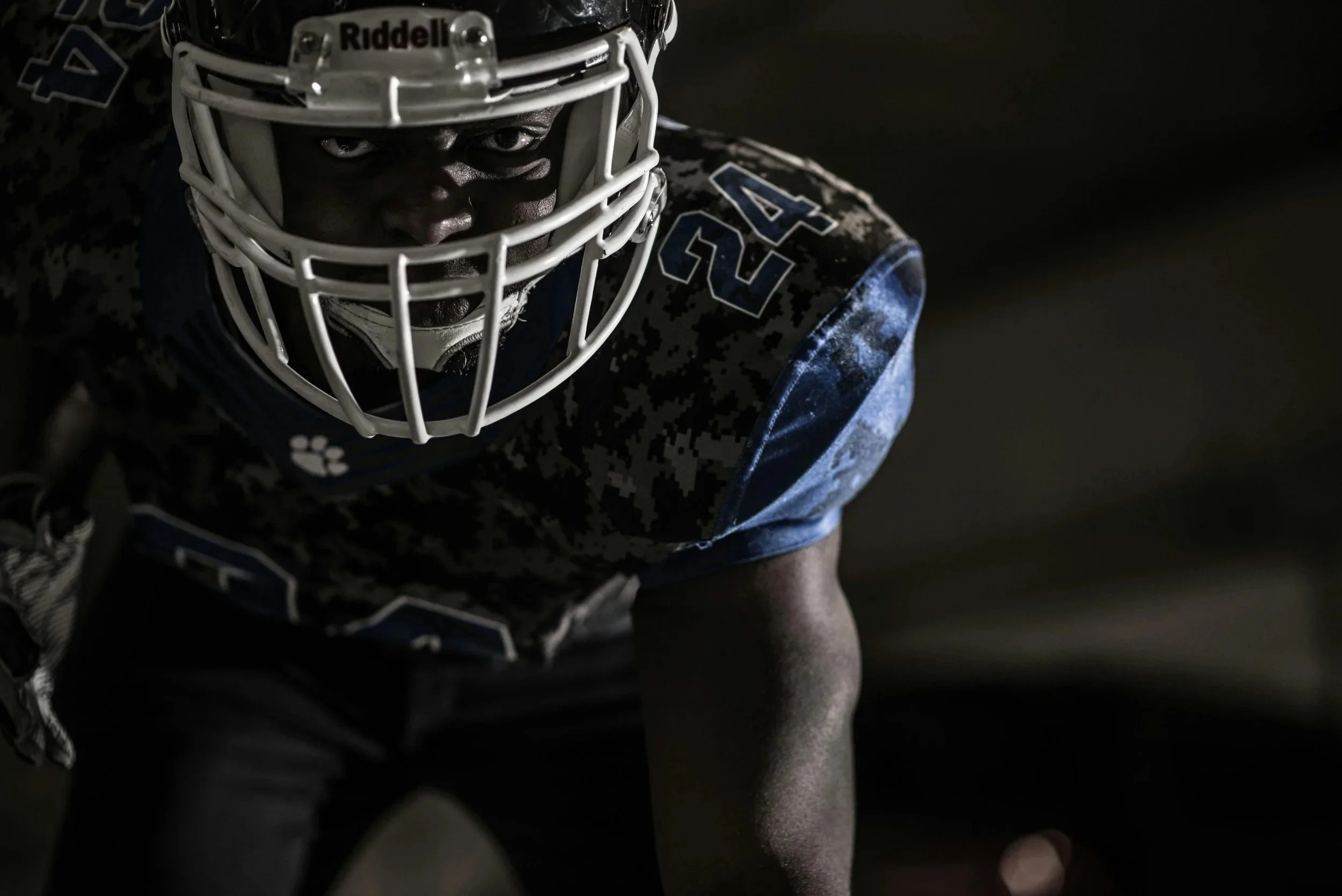 A football player crouched with a helmet, looking intensely at the camera, captured in a dimly lit environment.