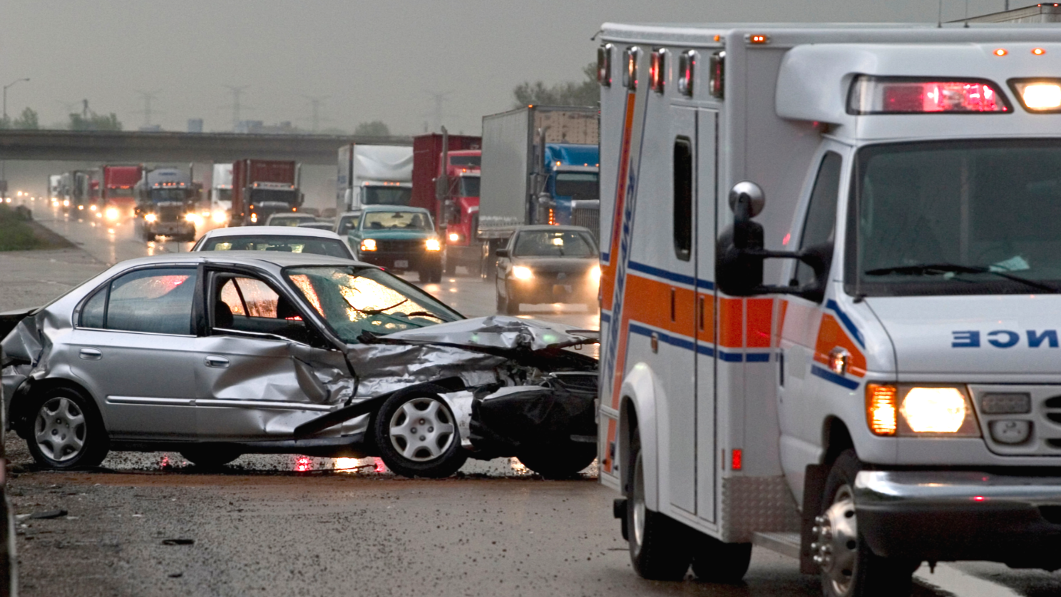A car accident scene on a highway with a severely damaged silver car in the foreground and an ambulance nearby. Traffic is backed up with multiple trucks and cars in the background, rainy weather with overcast skies. Houston