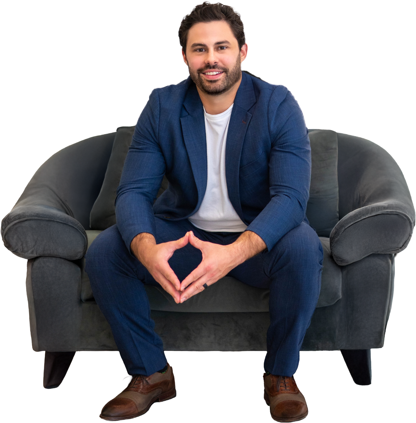 A man with dark hair and a beard sitting on a gray sofa, wearing a blue suit jacket, white shirt, blue pants, and brown shoes, smiling and sitting with his hands clasped in front of him.